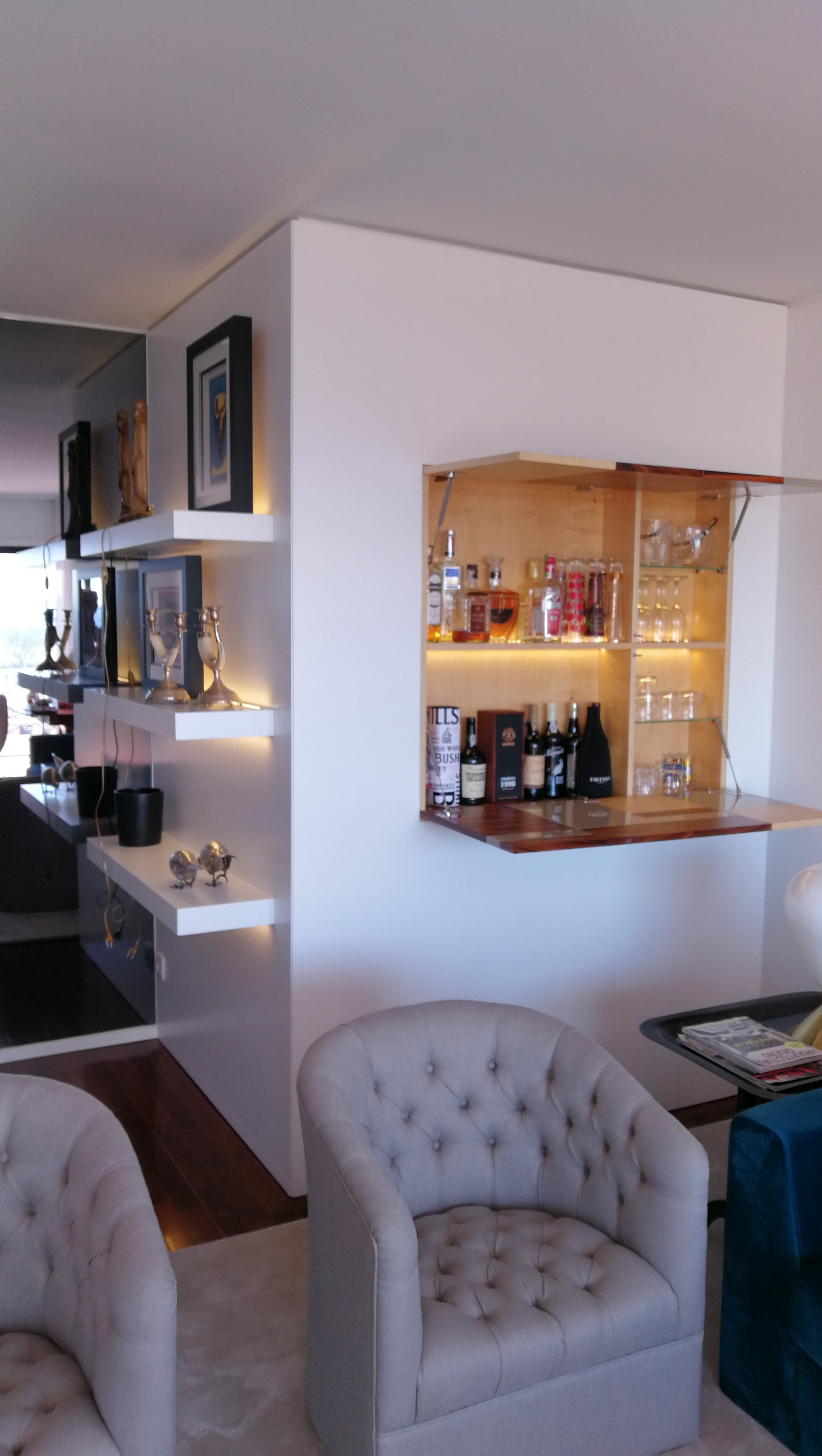 Sala com poltronas capitone, prateleiras iluminadas e bar embutido na parede com garrafas e copos de cristal