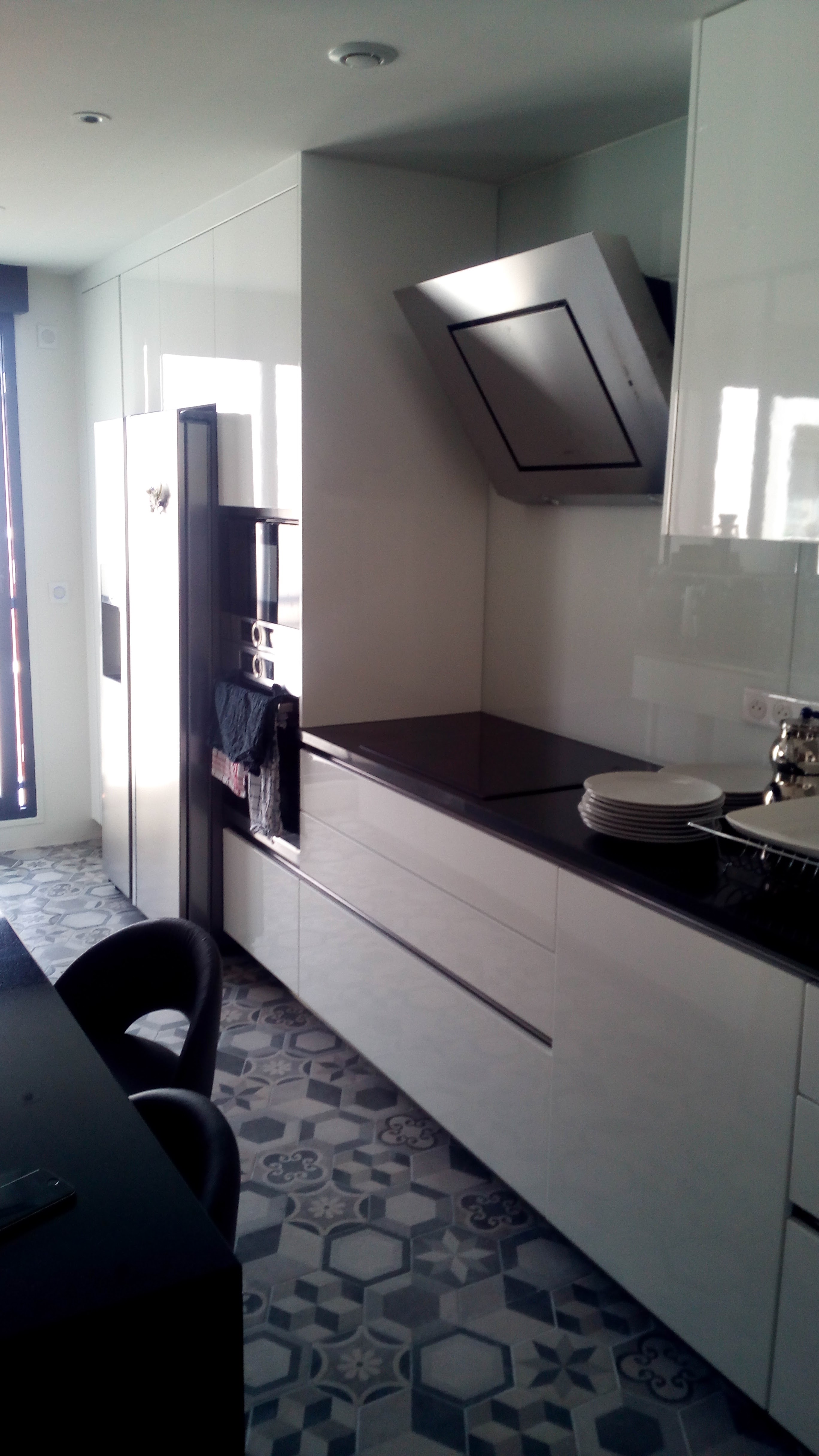 Cozinha com armarios brancos brilhantes, bancada em pedra escura, exaustor inox e pavimento em mosaico hidraulico