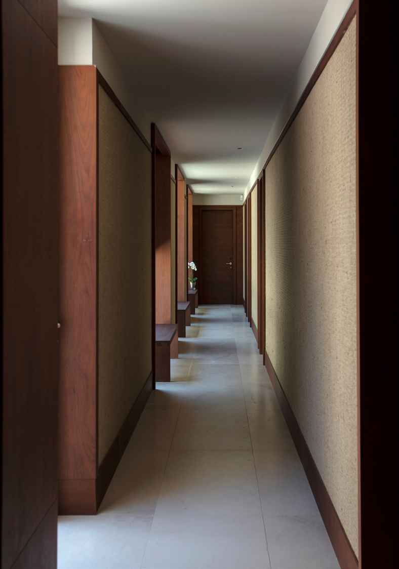 Corredor dos quartos com paredes revestidas em tecido natural, portas em madeira e iluminacao indireta