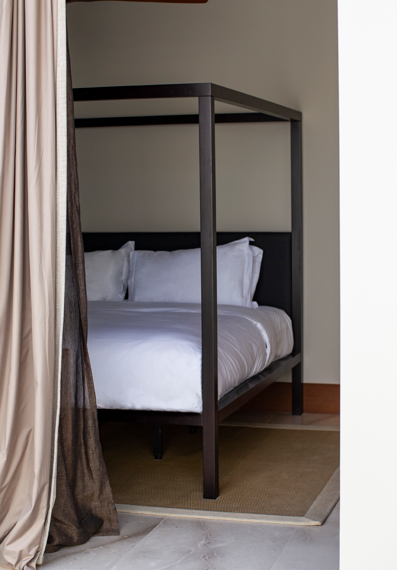 Quarto com cama de dossel em metal escuro, roupa de cama branca e cortinas em linho natural