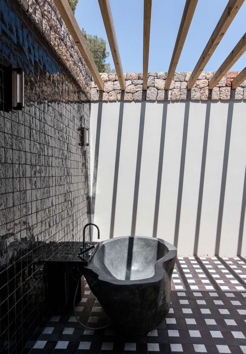 Duche exterior com banheira esculpida em pedra natural, revestimento em azulejo escuro e pergola em madeira