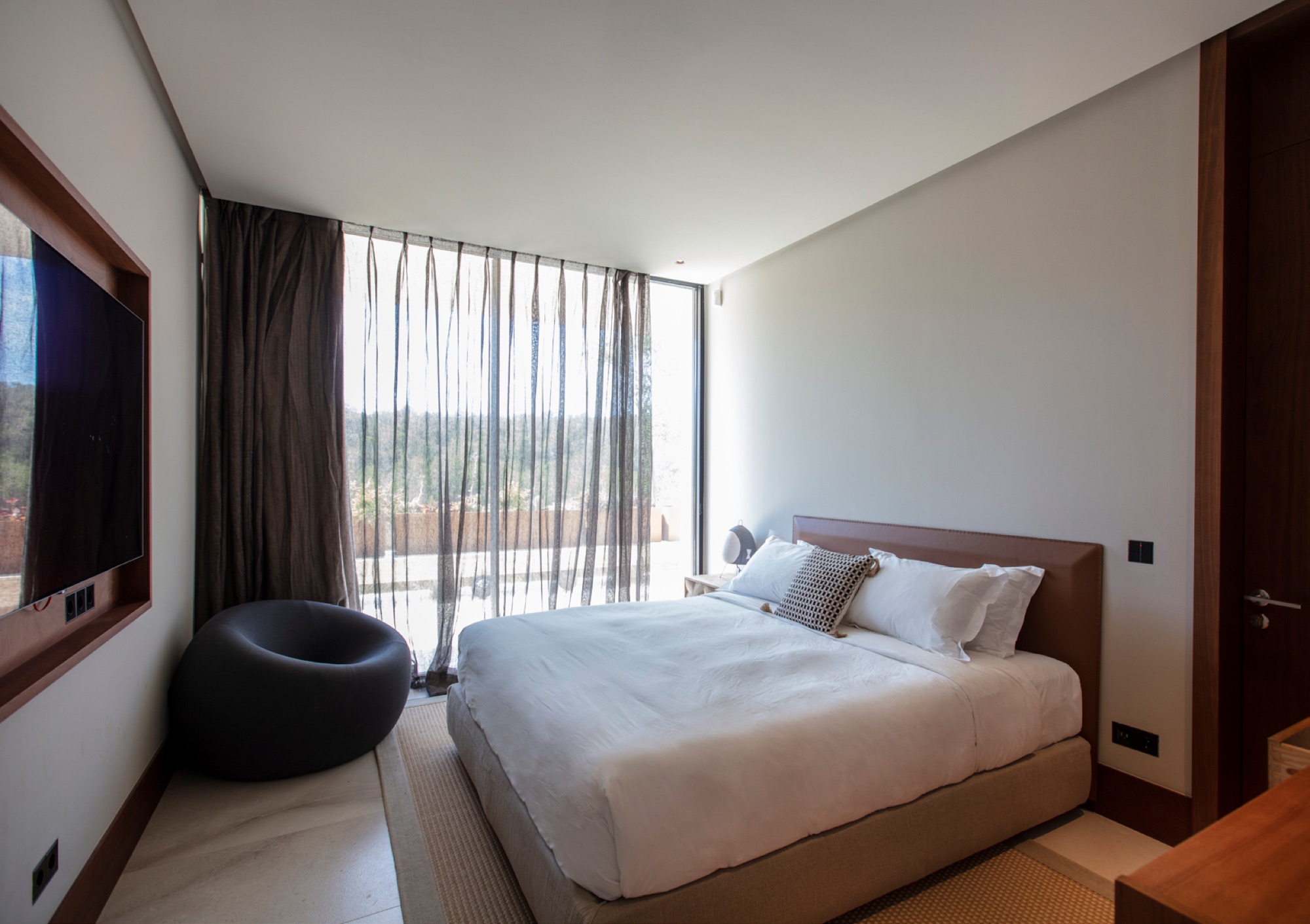 Quarto minimalista com cabeceira em madeira, roupa de cama branca, poltrona esferica e cortinas em linho