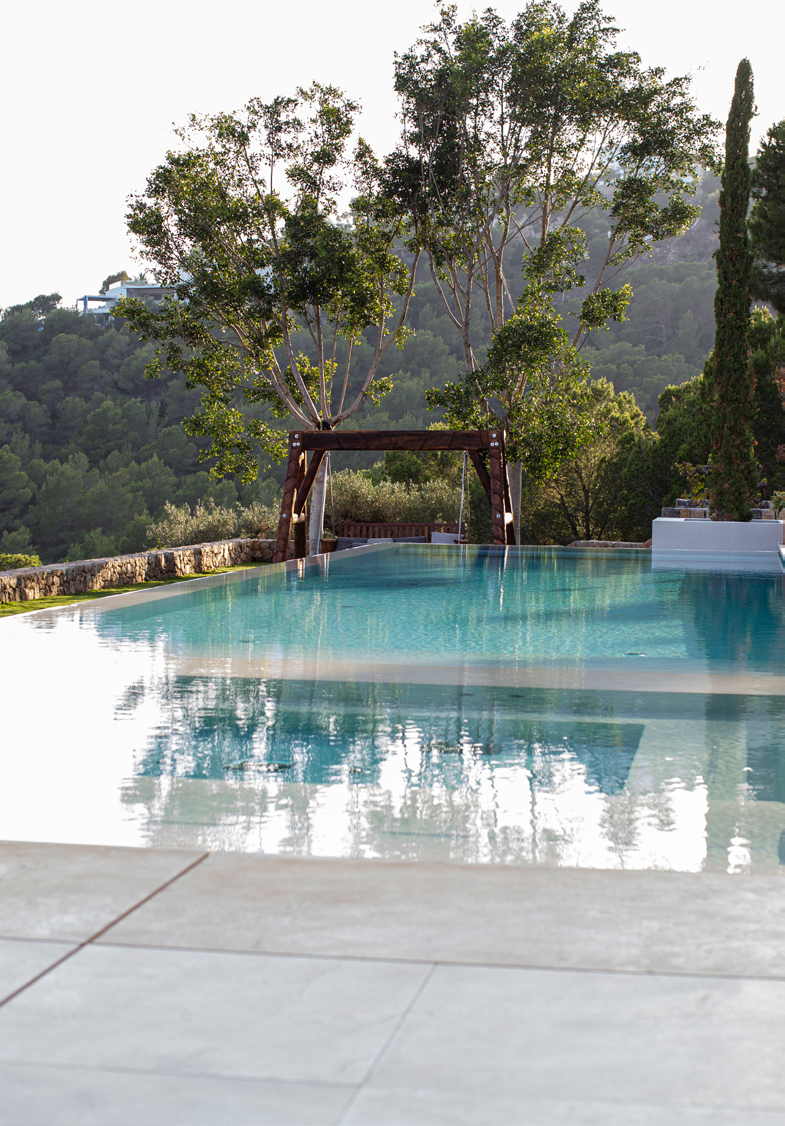 Piscina com pergola artesanal em madeira e cortinas, enquadrada pela vegetacao e colinas de Ibiza