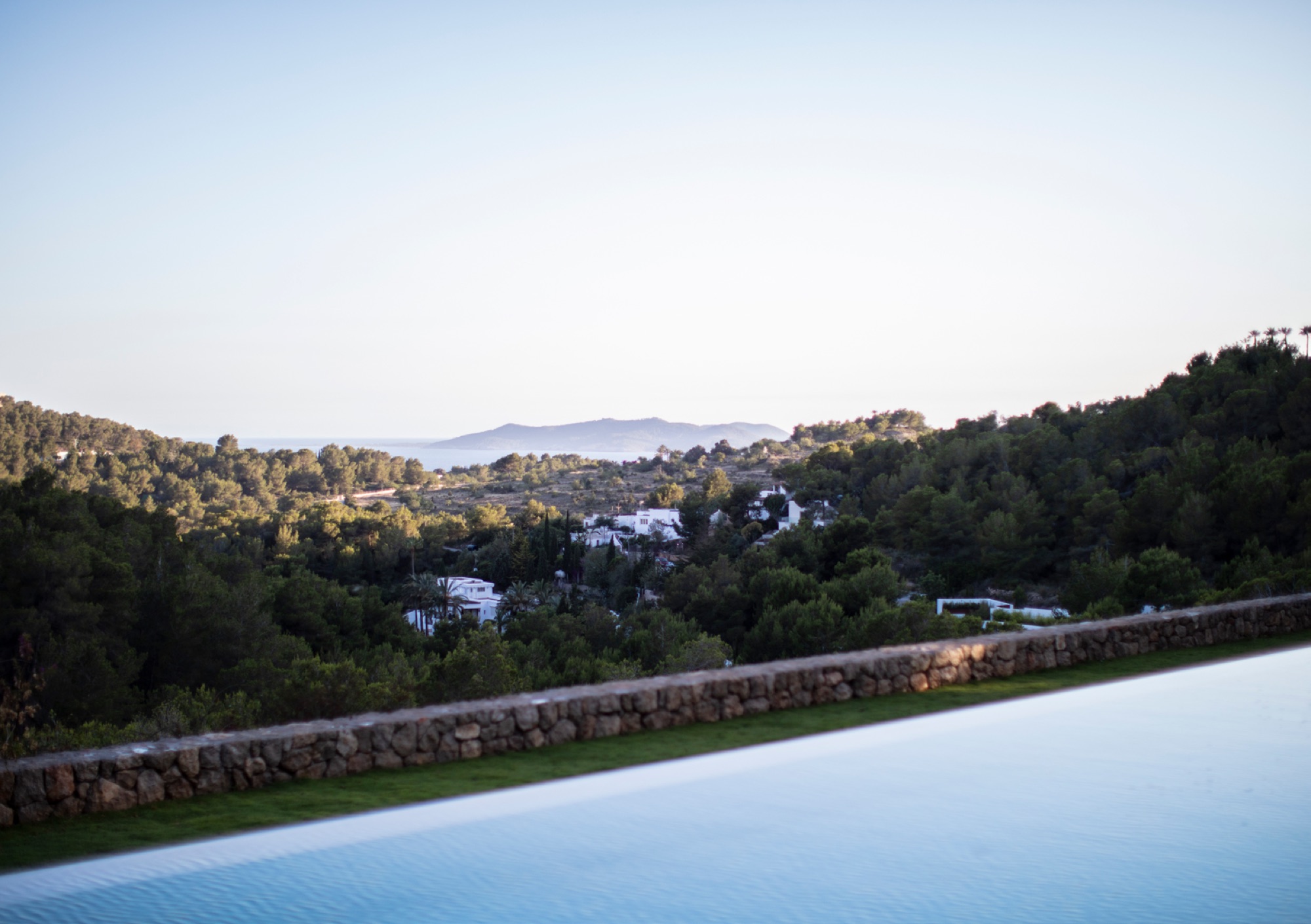 Piscina infinita ao anoitecer com muro em pedra natural e vista serena sobre as colinas e casas de Ibiza