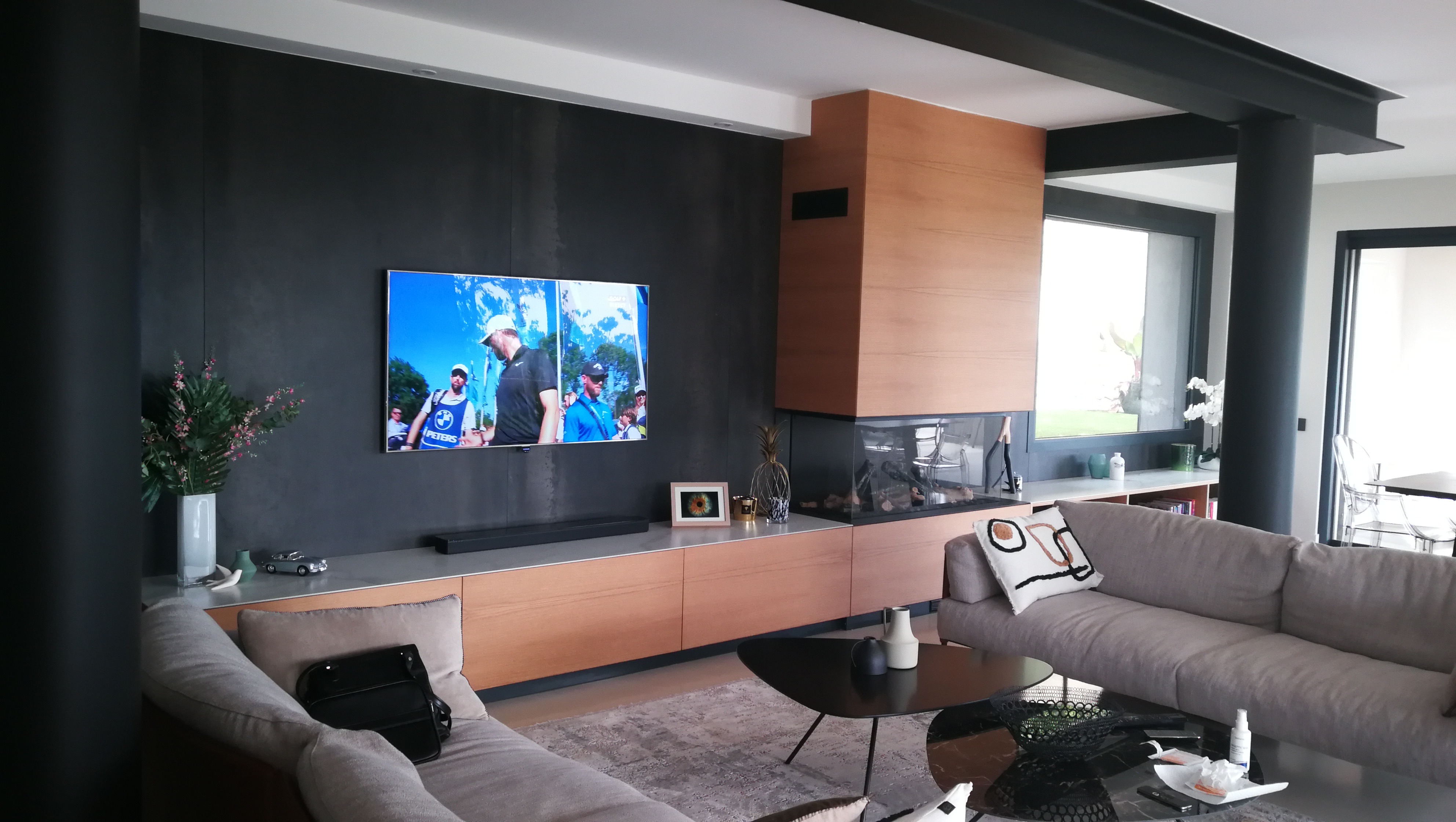 Sala com movel de parede em tons escuros e carvalho, lareira integrada e sofa cinza-bege